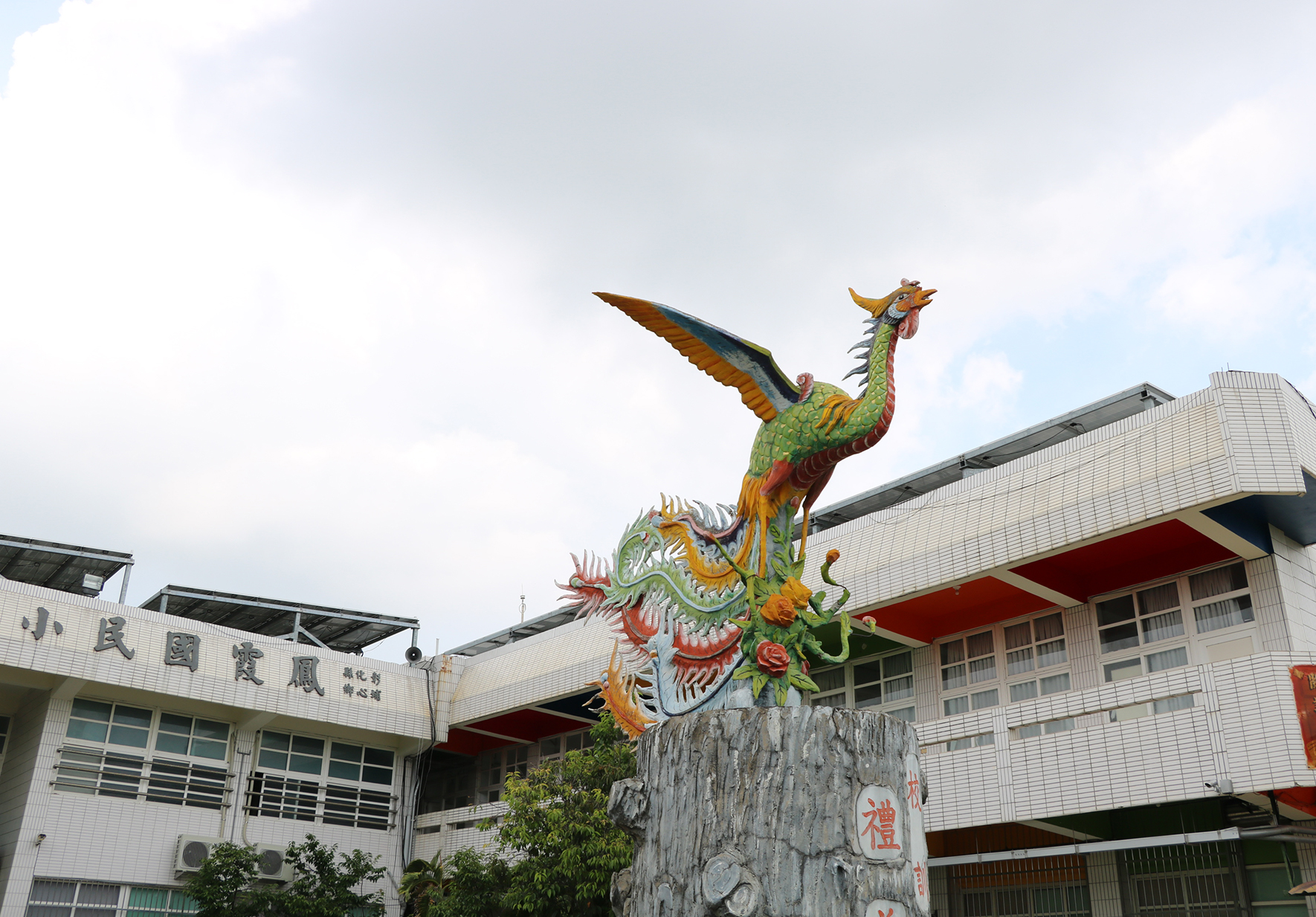 彩繪鳳凰雕像矗立在石基座上，展翅飛翔，身體繽紛多彩，包含綠、黃、紅等色彩。背景為現代校園建築，白色磁磚牆面搭配紅色設計元素。左側建築可見「孔子」字樣，顯示為教育機構。