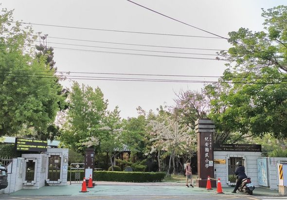 公園入口處，左右兩側各有一座黑色標誌牌，中間豎立紅色路錐，綠草地上有行人走動。背景是茂密的樹木和電線，天色陰沉。