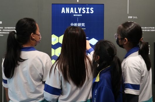 四名穿著統一校服的學生從背後觀看展示板。展示板標題為「ANALYSIS 分析研究」，以藍色背景搭配黃色幾何圖形設計。右側可見「STEP 2-1」文字及其他內容說明。