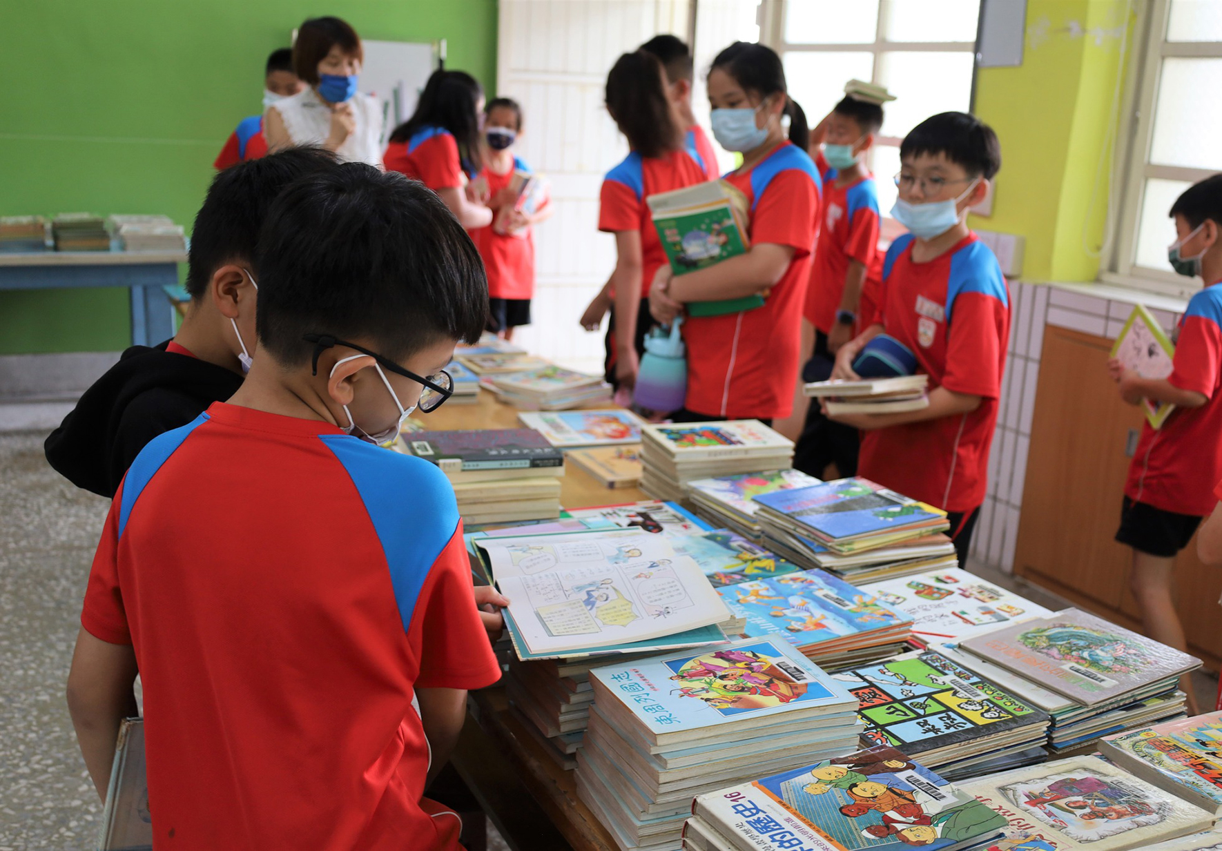 一群穿著紅藍色校服的小學生在教室內進行圖書館活動，學生們戴著口罩，圍繞著擺滿各式兒童書籍的桌子，正在挑選和閱讀圖書。教室背景為綠色牆面和明亮窗戶。
