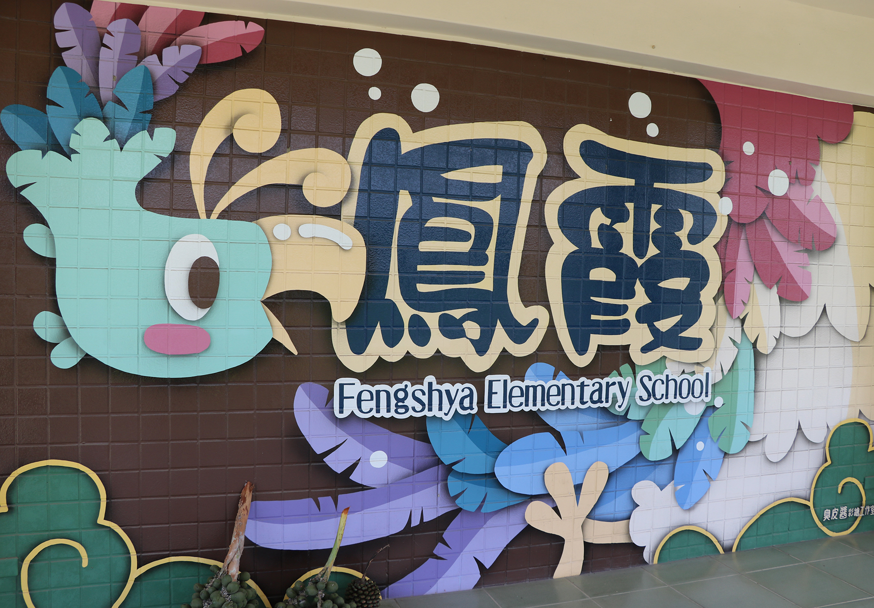 風沙國小校名標誌的彩色壁畫，以藍色、黃色、紫色和綠色為主。上方有中文校名，下方寫著「Fengshya Elementary School」英文校名。周圍裝飾著多彩紙質藝術花卉和仙人掌造型的校園牆面藝術。