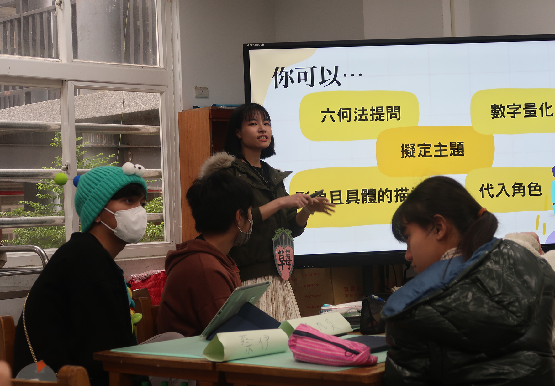 教室內，一位女性講師在觸控螢幕前為學生授課。螢幕顯示繁體中文教學內容，包括「六何法提問」、「擬定主題」等黃色文字框。三位戴口罩的學生坐在課桌前聽講，桌上有筆記本和文具。背景是教室窗戶和書櫃。