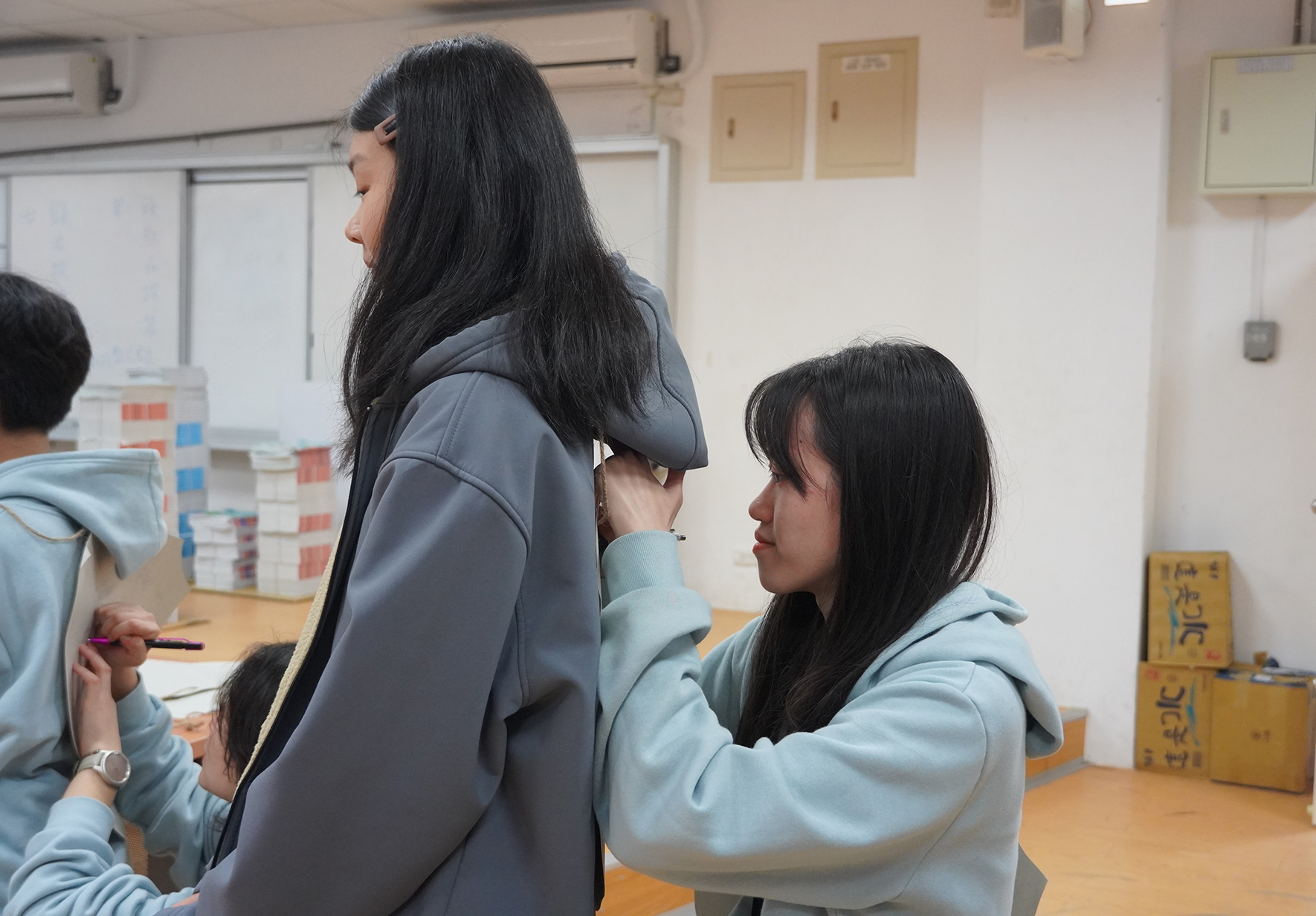 教室內，三名穿著灰藍色連帽上衣的年輕女性正在進行小組討論。站立的女性與坐著的女性面對面交談，背景可見白板、教學用具和日文標示牌。