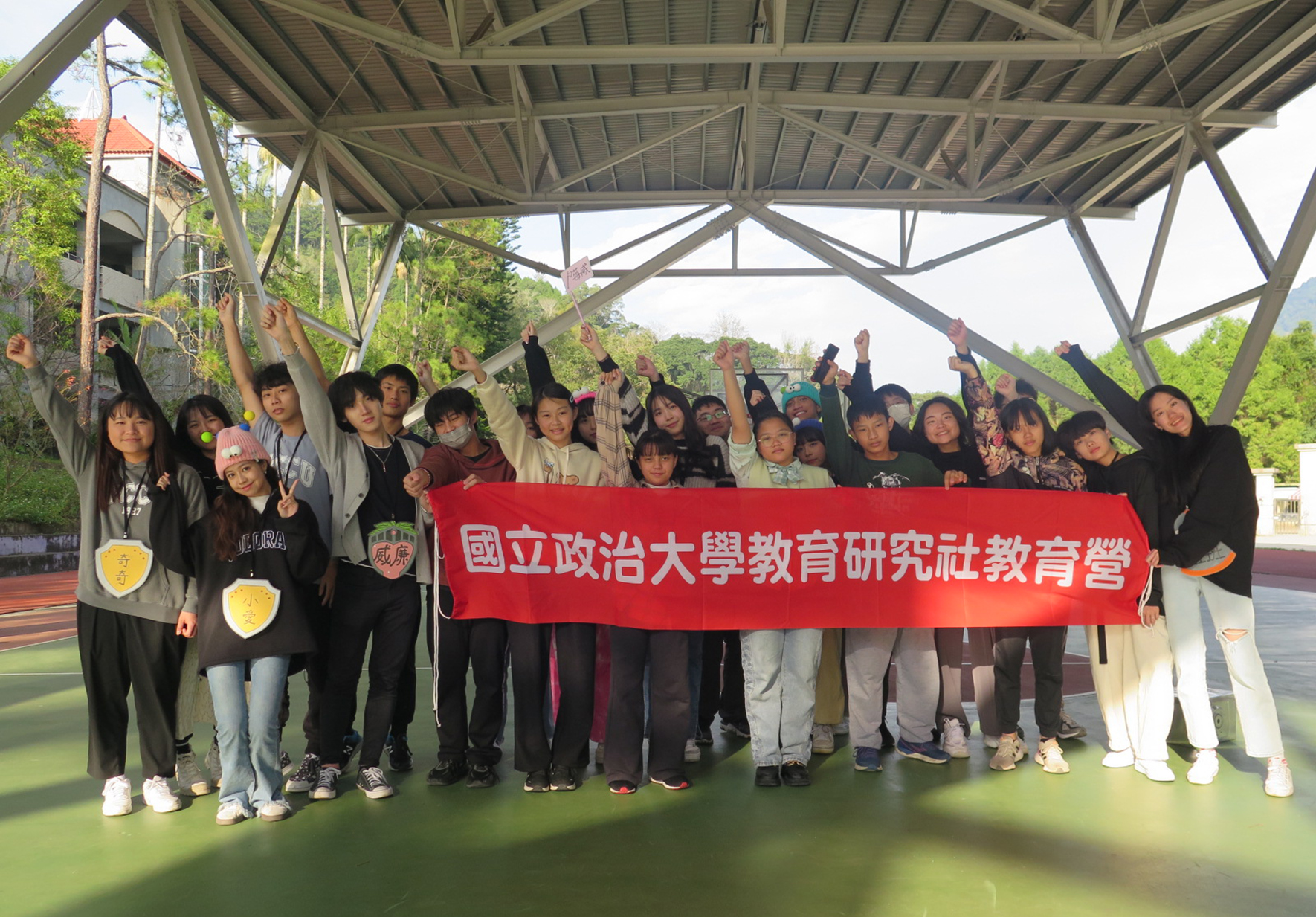 國立政治大學教育研究社教育營的參與者在室外遮陽棚下集體合影，眾人舉起雙手做勝利手勢，中央展示紅色橫幅，上面寫著「國立政治大學教育研究社教育營」。
