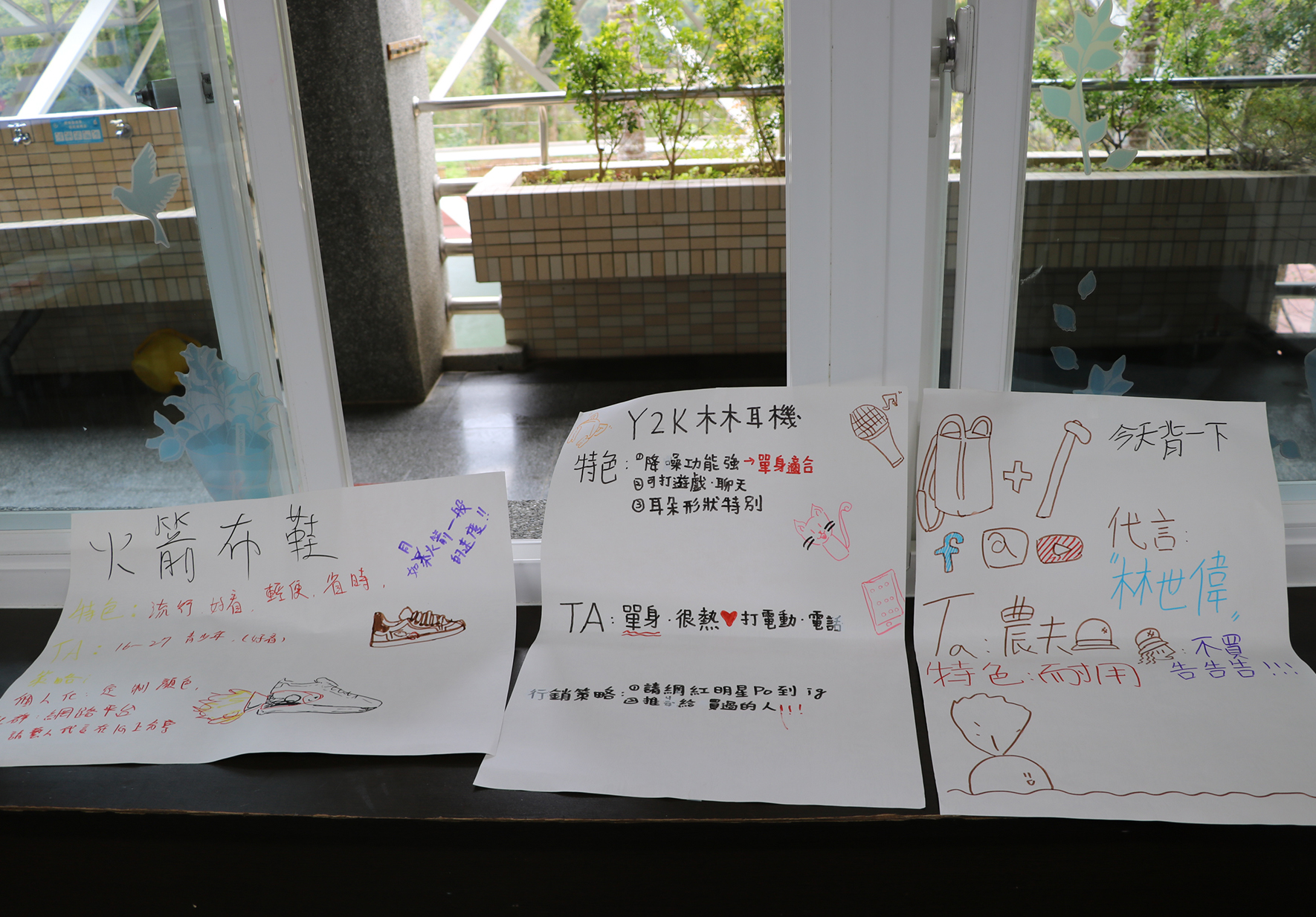 窗邊展示台上，三張手寫海報並排放置，內容以繁體中文書寫，介紹不同產品或主題。左側海報談論水前折疊鞋，中間為Y2K木木耳機產品介紹，右側為秋背包代言商品。各海報搭配彩色手繪插圖和圖示。背景可見落地窗、綠植和室外環境。