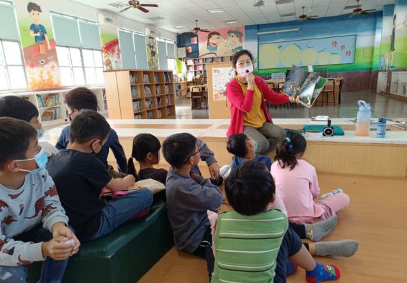 小學圖書館內，一位女老師穿著紅色外套，站在講臺前向坐成一圈的學童們展示繪本故事書。教室牆面貼滿彩色教學海報和學生作品，書架擺放著多本圖書。這是一場生動的兒童故事講述活動。