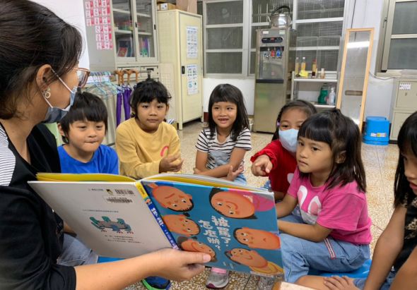 教室內，一位女性教師向圍坐的小學生展示繪本。書上有橙色熊貓圖案。約六名學生專注聆聽，背景有書櫃和教室佈置。進行中文閱讀教學活動。