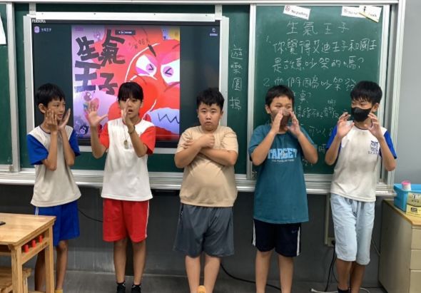 五位小學生在教室裡站成一排，背靠黑板和螢幕，做出思考的手勢。教室背景有中文字跡和紅色藝術作品展示，顯示是課堂學習活動或小組討論的情境。