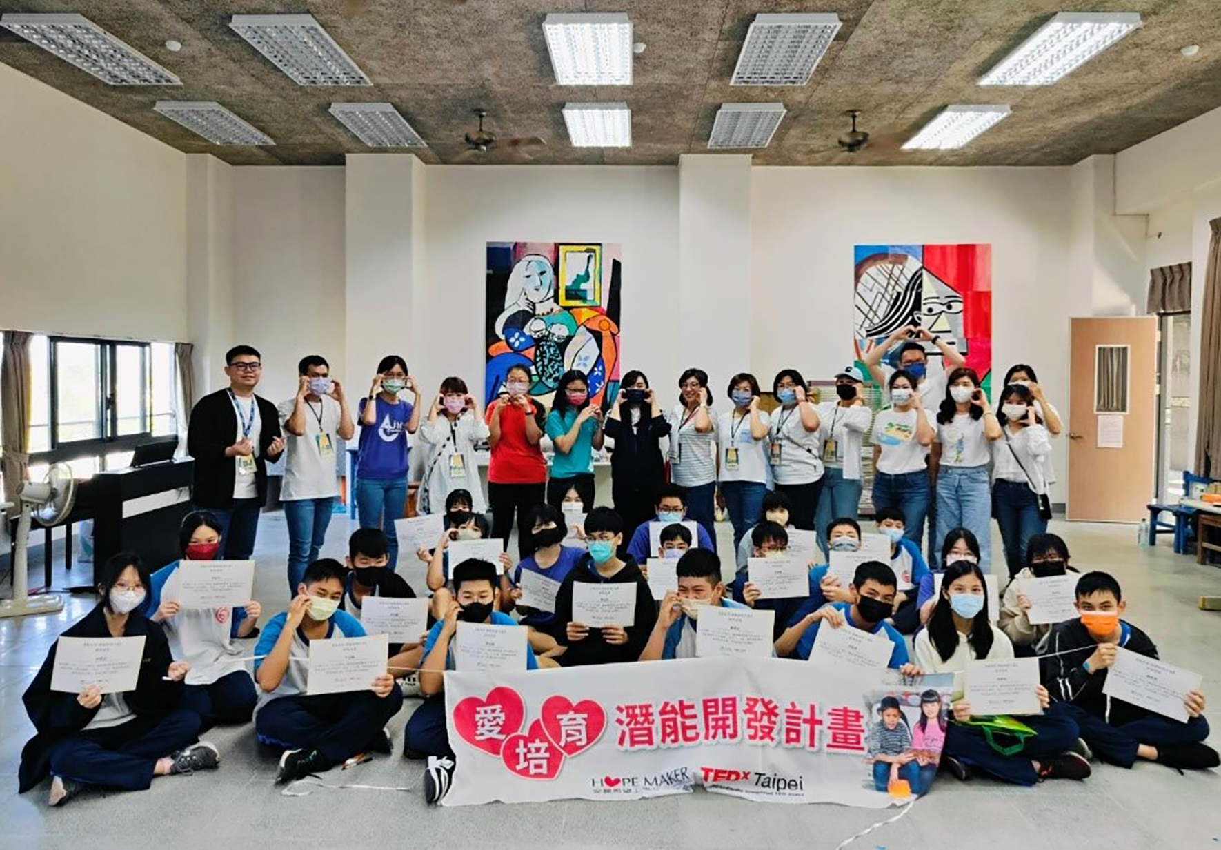 一群戴著口罩的學生在室內活動現場合影，前排坐著，後排站著，均舉起證書。橫幅寫著「愛培育潛能開發計畫」和「TEDxTaipei」的標誌。牆上掛著彩色藝術作品，天花板有多盞照明燈。