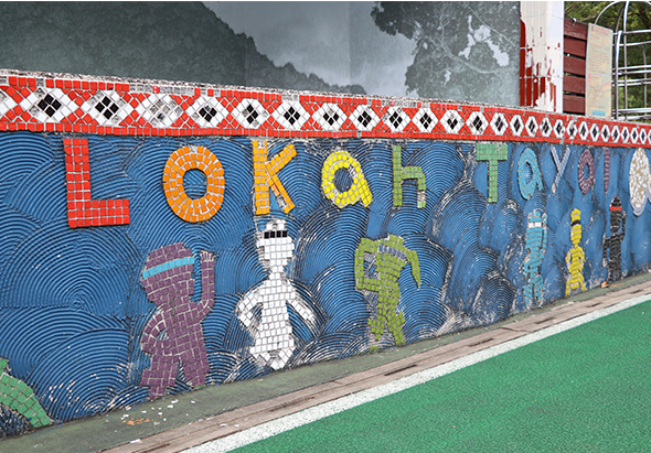 彩色壁畫展示「Lokaytaya」文字，藍色背景配紅白幾何邊框裝飾，繪有傳統建築、人物和文化圖案，山景映襯背景，前方為綠色地面。