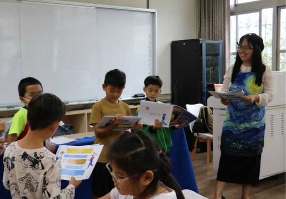教室內，女教師站在講台旁向坐著的七名學生進行教學。學生手持教材和學習資料，教室背景有白板和教學設備。這是一堂小班制課堂教學活動。