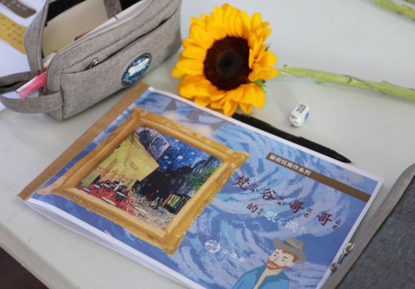 白色桌面上擺放著一本藍色封面的書籍和一幅金色相框的梵谷名畫《夜間咖啡館》複製品。旁邊放著一朵黃色向日葵、灰色布質背包和白色USB隨身碟，展示梵谷藝術相關的書籍和紀念品。