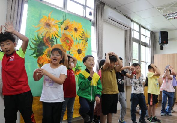 一群小學生在教室內進行藝術活動，身後是一幅描繪向日葵的大型壁畫。孩子們穿著不同顏色的衣服，擺出歡樂的姿勢，有些舉起雙手，有些做出開心的表情，展現出活潑的課堂氛圍。