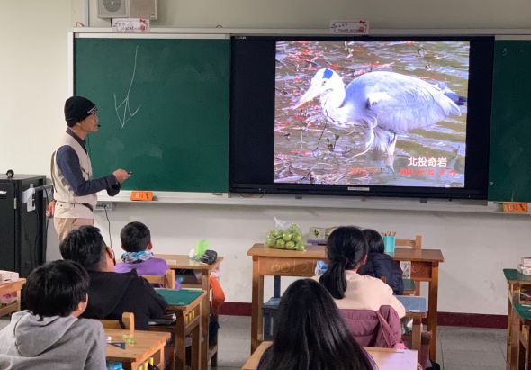 教室裡，教師站在講臺旁向學生授課，大螢幕上顯示一隻白色水鳥（北設當岩）在水邊的影像。多名學生坐在課桌前認真聽講，進行自然生物教學活動。