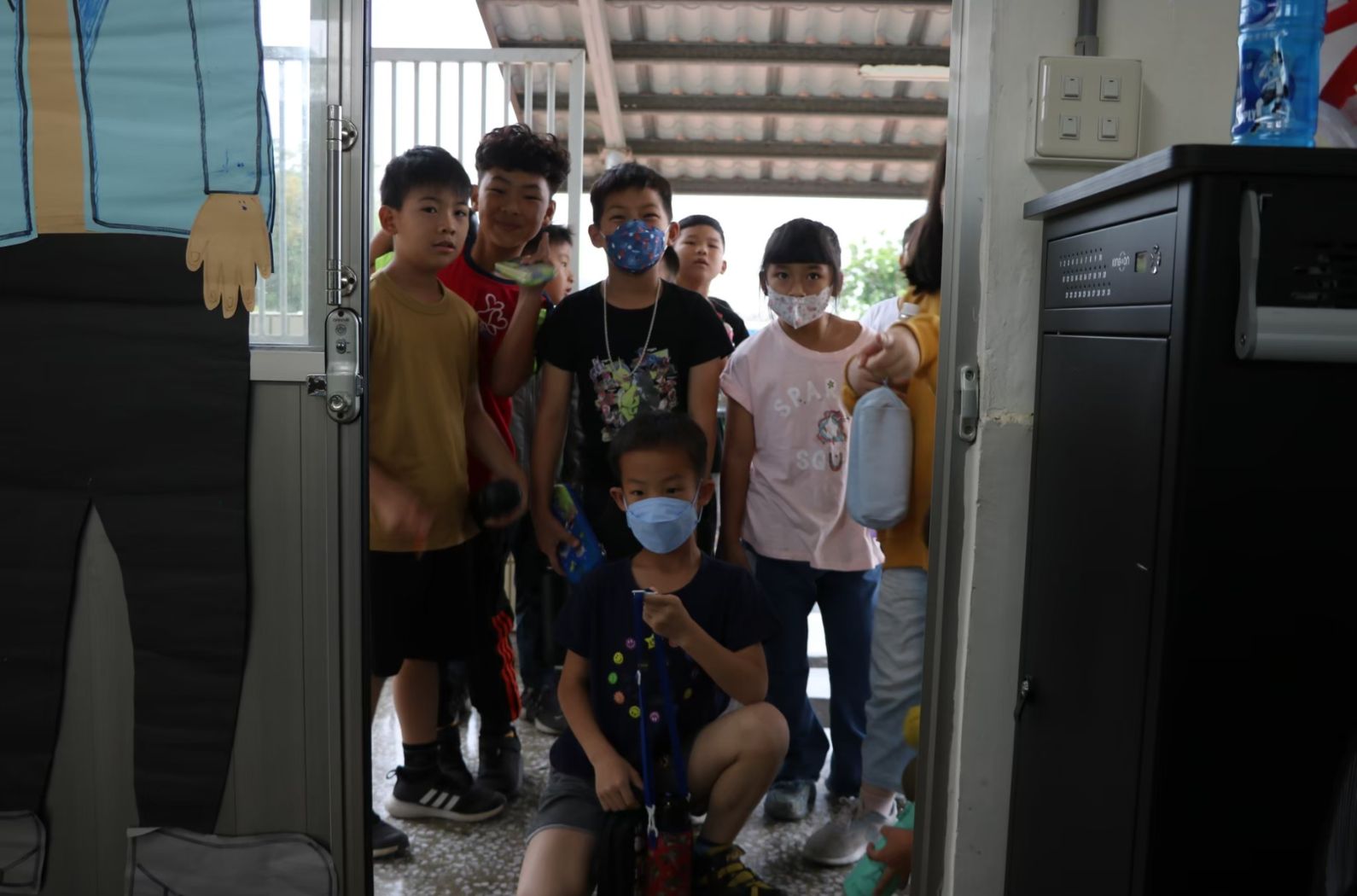 一群兒童在室內聚集在開放的門口前，其中多數戴著口罩。他們穿著各色T恤，站在簡樸的教室環境中。背景可見樓梯和戶外景象。這是一張學校或教育機構的集體活動照片。