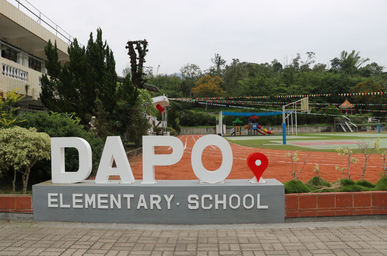 達波小學校園入口標誌。白色大字「DAPO」搭配紅色地點符號，下方灰色底座標示「ELEMENTARY SCHOOL」。背景可見紅色跑道、綠色草坪、運動設施及校舍建築，彩旗裝飾校園。