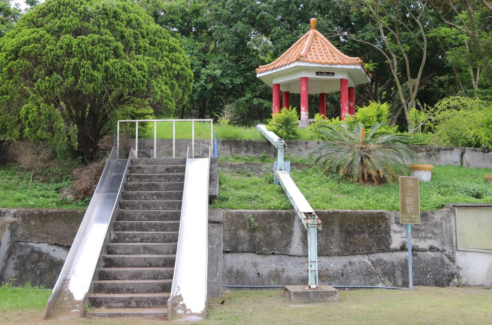 公園內有石階樓梯和白色滑梯供遊客上下，階梯兩側設有玻璃護欄。上方平台有紅柱子支撐、橙色琉璃瓦頂的亭子建築。周圍被茂密的綠樹和草地包圍，右側有棕櫚樹和標示牌。
