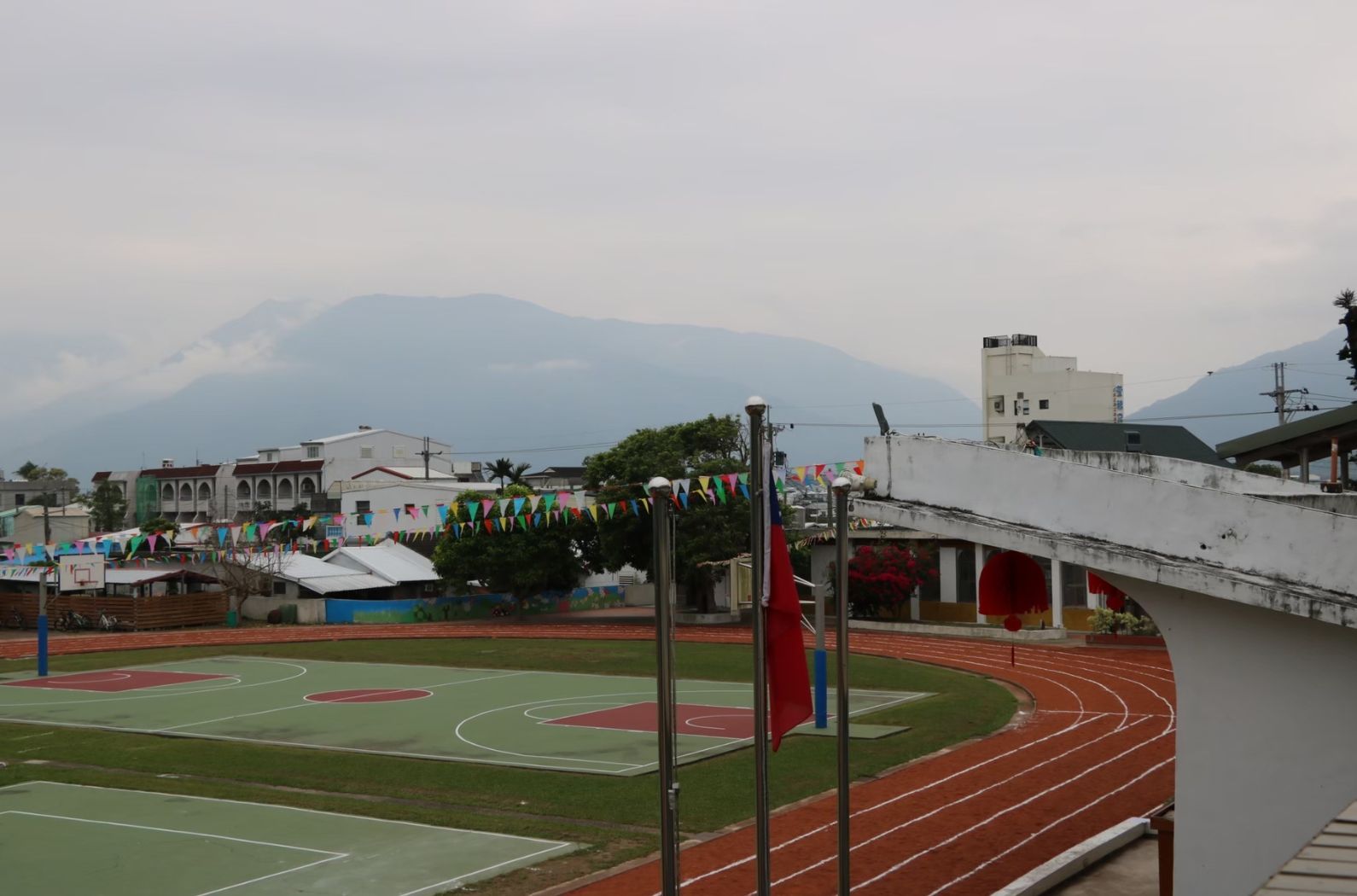 學校運動場景，紅色跑道環繞綠色籃球場，白色帳篷和彩色旗幟裝飾校園，背景為壯麗的大山，展現校慶或運動會的熱鬧氛圍。