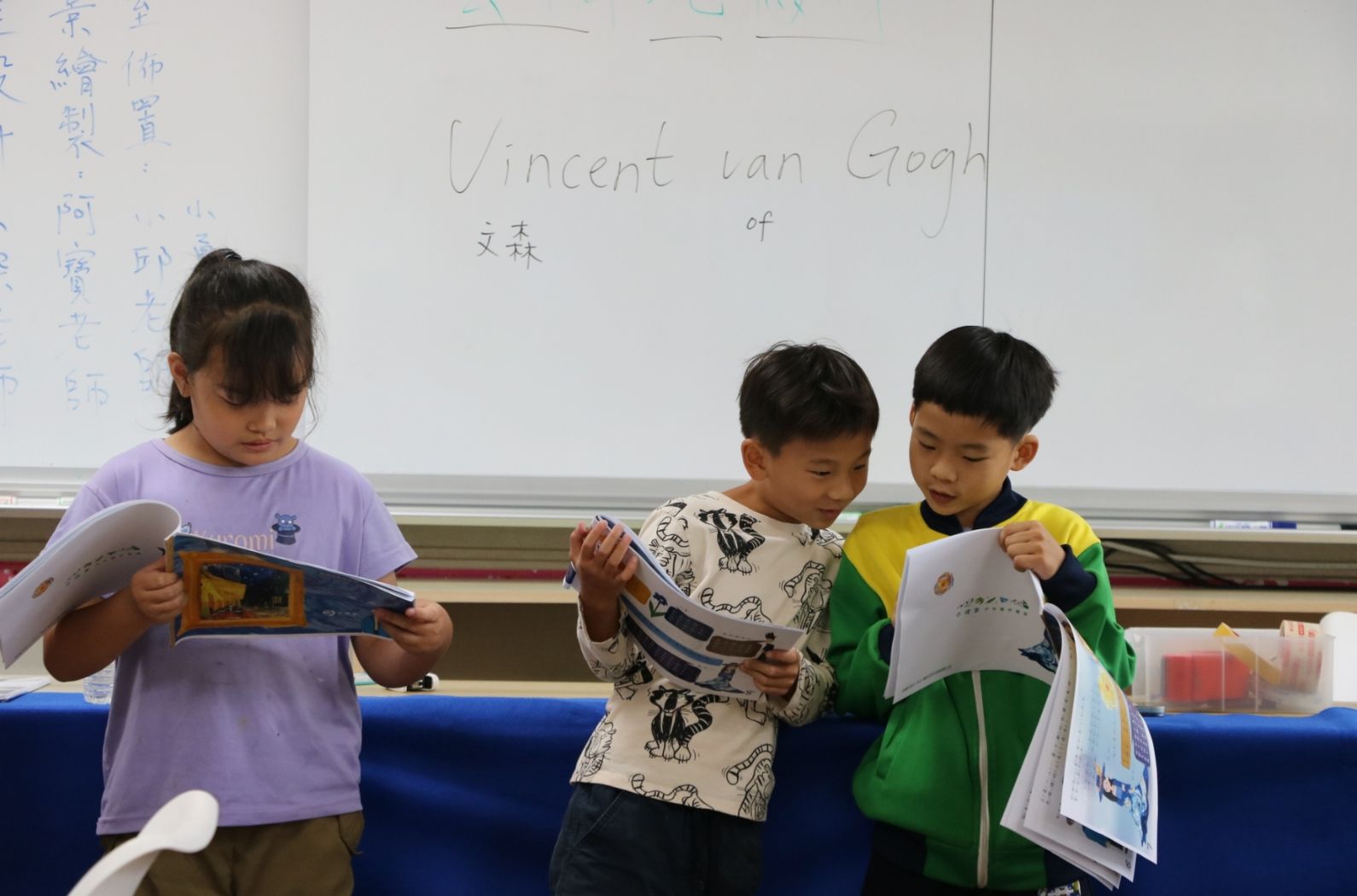 三位小學生在教室內閱讀書籍。背景白板上寫著「Vincent Ian Gogh」及中文字，左側站著穿紫色上衣的女童，中間穿花紋衣的男童，右側穿綠色上衣的男童，三人專注低頭閱讀各自手中的書冊。