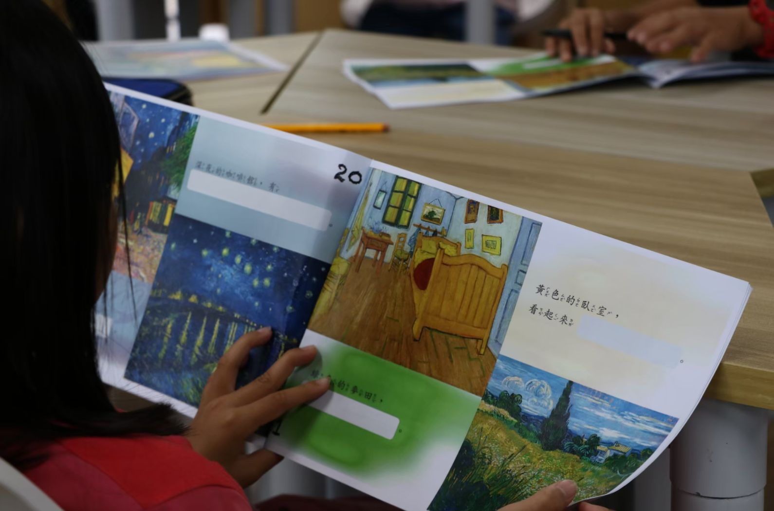 一位學童在課堂上手持打開的繪本，展示第20頁內容。繪本展示梵谷的臥室畫作及其他經典藝術作品，並附有中文文字說明。木製課桌上散放著多本藝術相關繪本和鉛筆，其他學童在背景中閱讀。