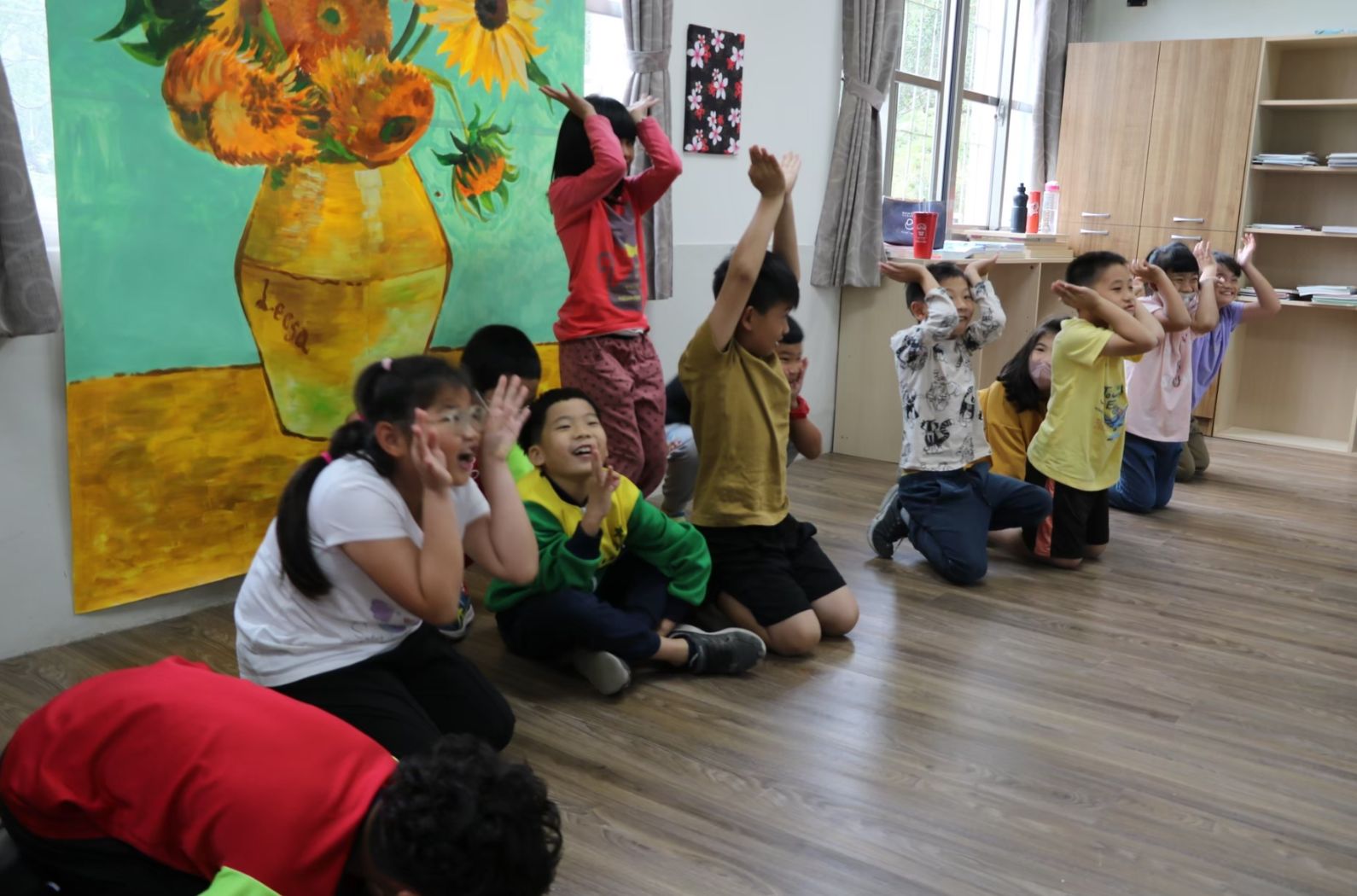 一群小學生在教室內跪坐地板上，舉起雙手做動作，進行互動遊戲或教學活動。教室牆上掛著梵谷《向日葵》畫作，窗邊有木製書架和教學用品。