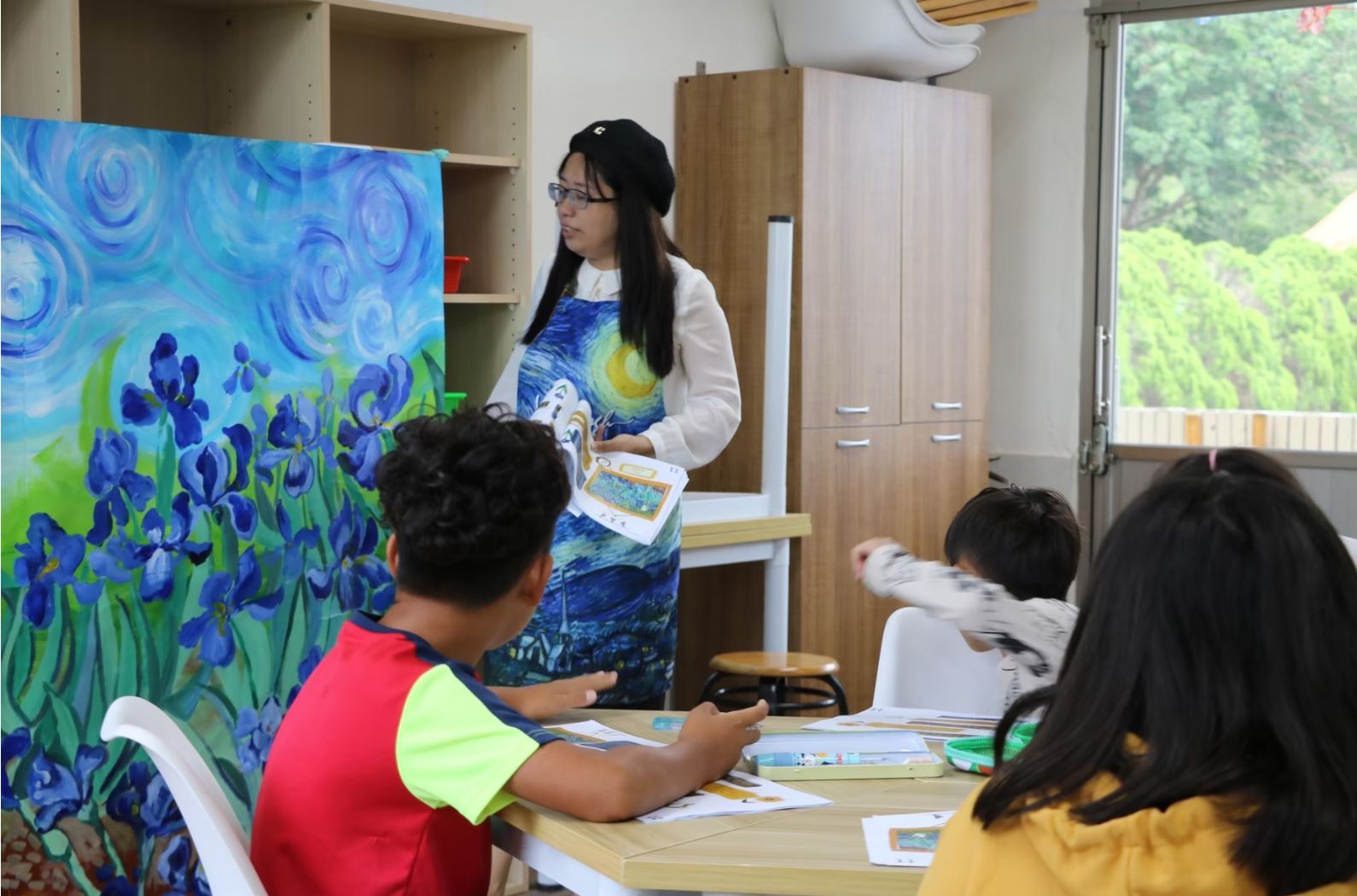 藝術課堂上，女教師穿著印有梵谷名畫的服裝，向學生展示藍色鳶尾花主題的畫作。三位學生坐在桌前認真聽講，桌上放有畫紙和繪畫用具，教室背景為木質家具和綠色窗外景致。