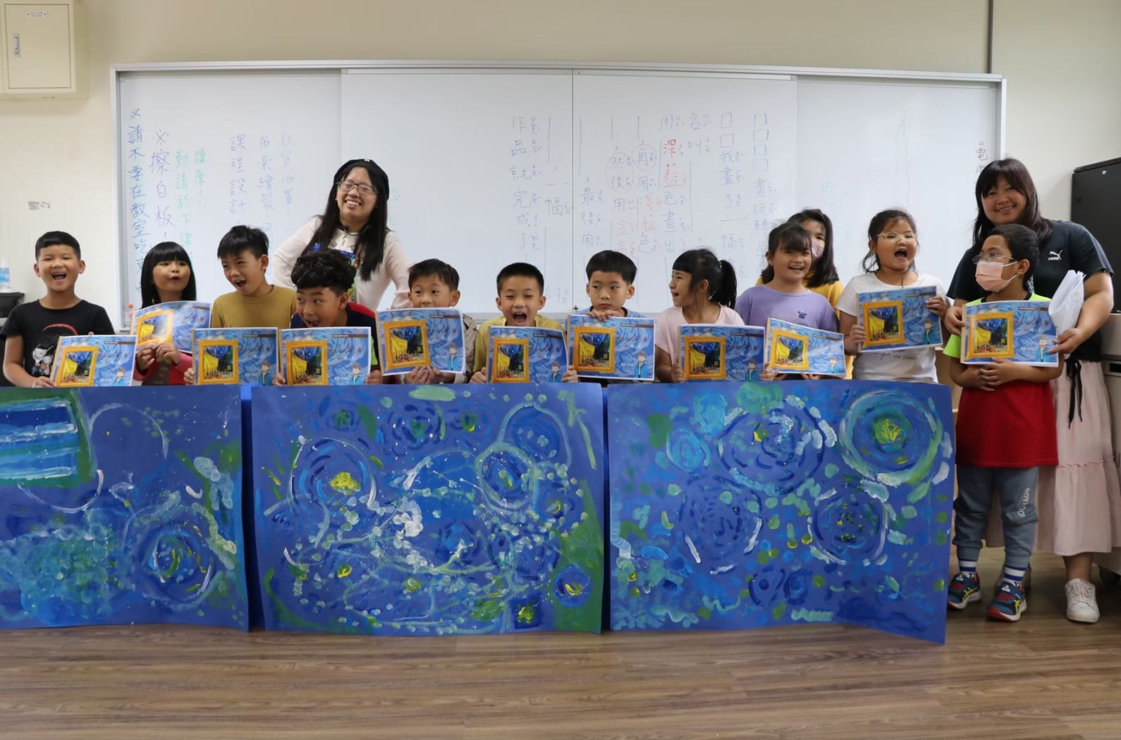 一群小學生在教室內展示美術作品。他們手持繪有黃色圖案的藍色作品，身後是大型藍綠色集體繪畫，背景有白板和教師。孩子們開心地微笑，呈現藝術學習成果。