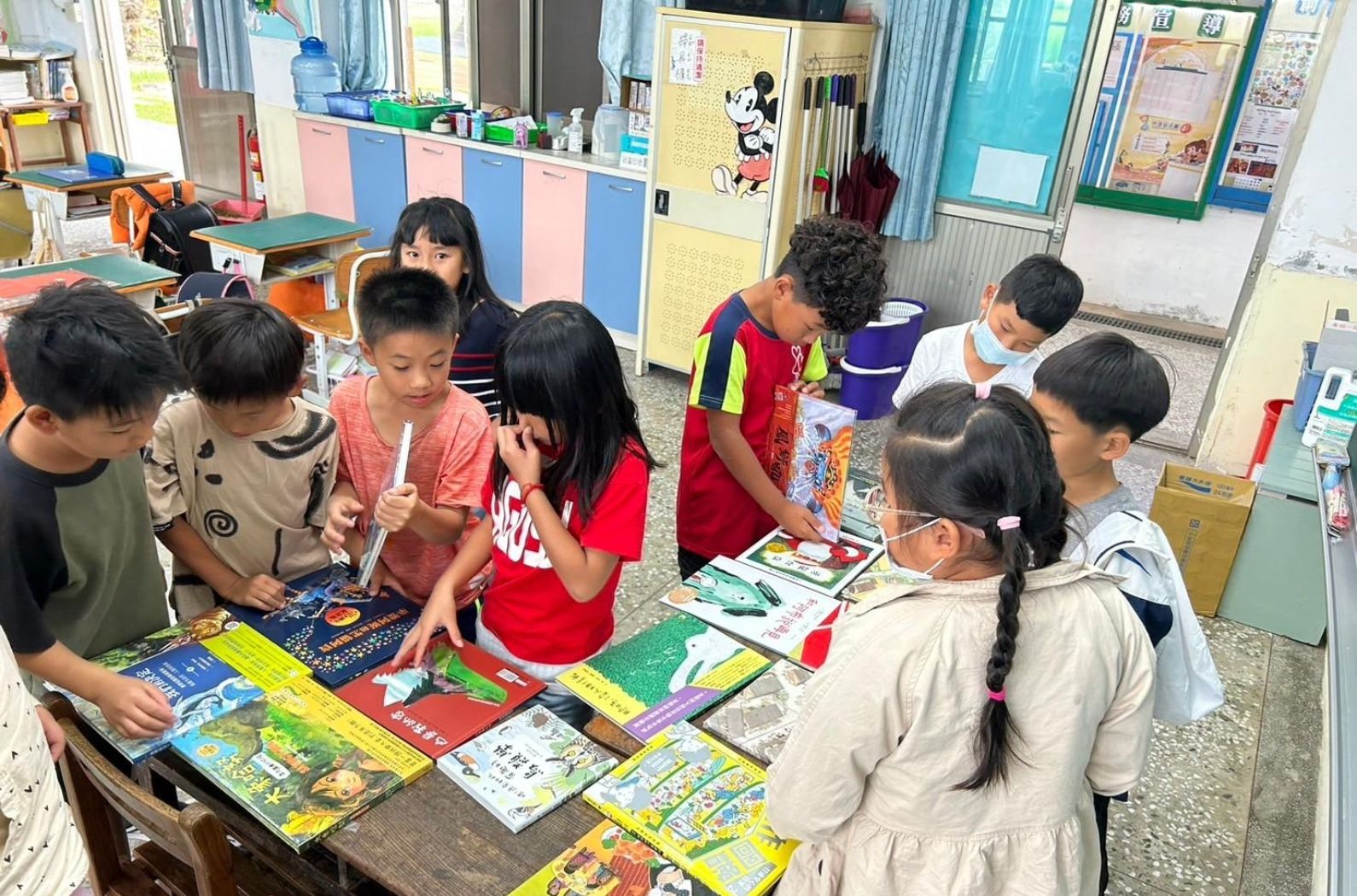 小學教室內，約十名學童圍聚在一張桌子周圍，認真地瀏覽和選擇擺放在桌上的各色兒童書籍。背景可見粉藍色的儲物櫃、黃色佈告欄和教室布置，營造出溫馨的閱讀推廣活動氛圍。