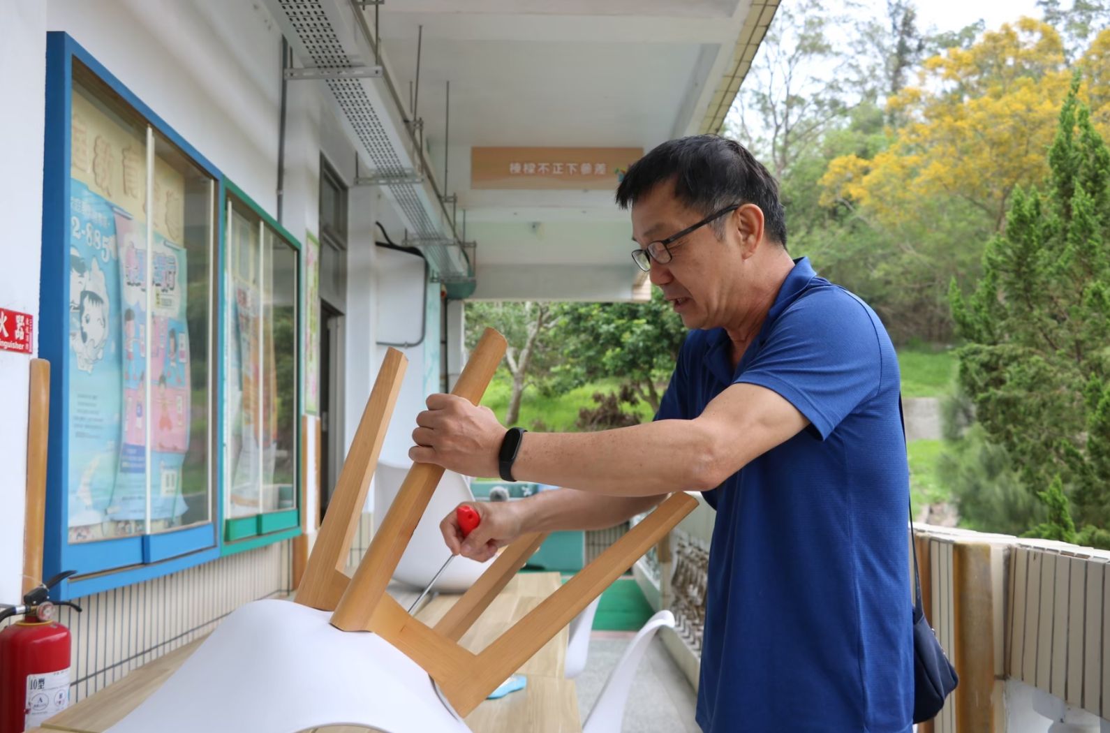 一名穿著藍色短袖上衣、戴眼鏡的男性，在室內走廊上組裝木製的A字型架子。左側牆上掛著藍色邊框的公告欄。背景可見窗外綠色樹木景致。走廊內還有紅色滅火器和其他設施。