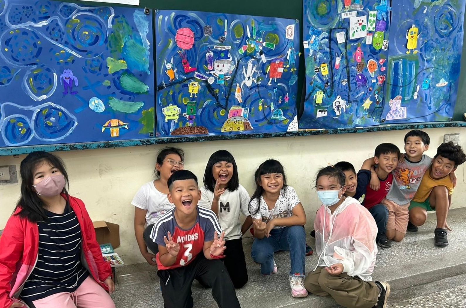 一群小學生在教室裡蹲坐成一排，面帶笑容。身後牆上掛著藍色主題的太空宇宙主題壁畫，繪有行星、太空船和卡通角色。部分學生戴著口罩，展現歡樂的課堂氛圍。
