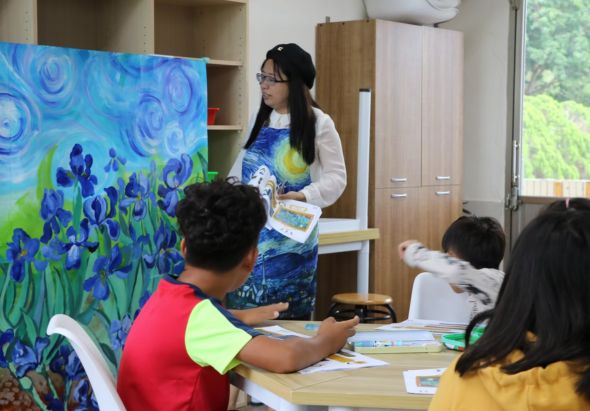 教室內，女性教師站在展示梵谷「星夜」風格的藍紫色花卉畫作前，向坐著的三位學生講解。學生桌上放有美術用具和教材，認真聽講和觀看藝術作品。