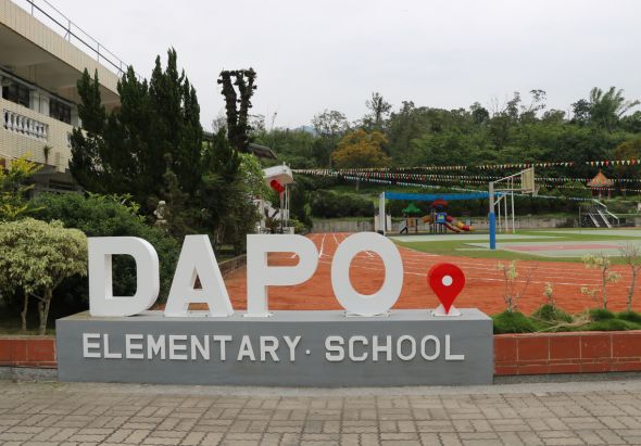 達坡小學入口標誌，白色大字母「DAPO」配紅色定位符號，下方灰色底板寫著「ELEMENTARY SCHOOL」。背景為校園操場、遊樂設施、綠樹和校舍建築。