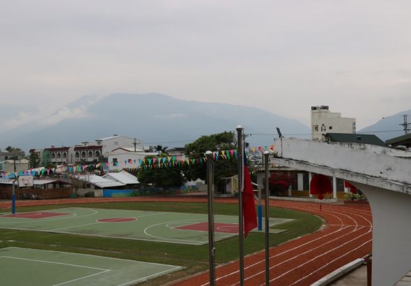 運動場景：紅色跑道和綠色籃球場地，背景有校園建築、帳篷和活動設置。遠處山巒環繞，天色陰沈。場地上方裝有排球網，顯示這是學校或社區運動會的舉辦地點。