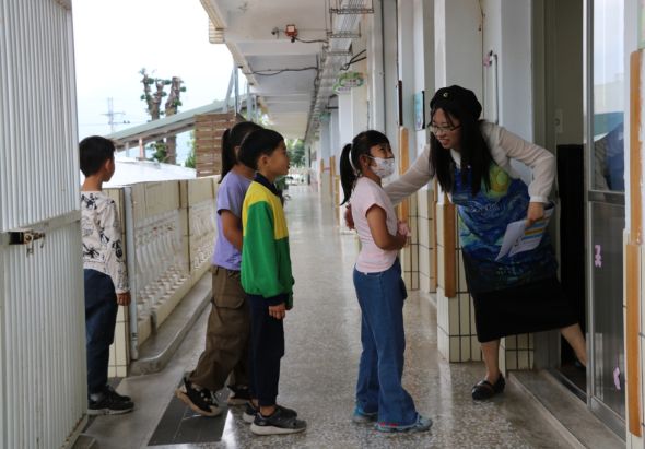 在學校走廊裡，一位女性教師與四名小學生互動。教師穿著藍色背心，正在與學生交談。學生們穿著各色校服，在走廊兩側站立。背景可見教室門窗和走廊設施。