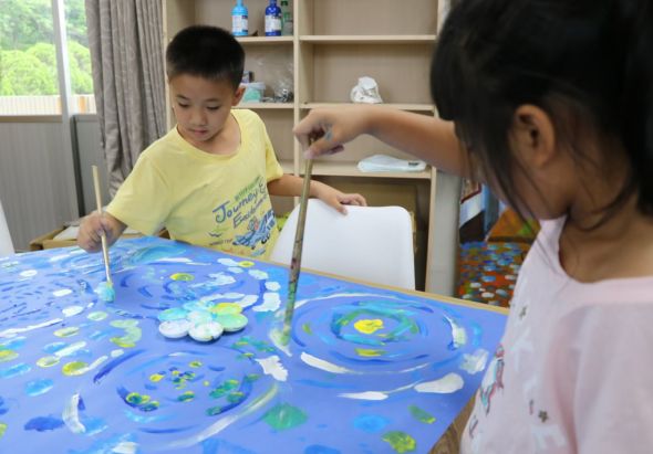 兩名小朋友在室內桌子上進行美術創作，他們用筆在藍色底的畫紙上繪畫，圖案中有螺旋紋、星星和各色圖樣。背景是書櫃、窗戶和灰色窗簾。