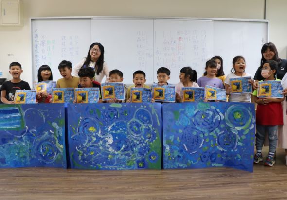 教室內，一位女教師與約十多位小學生站在三幅藍色主題的大型壁畫前展示作品。學生們手持黃色封面的書籍，壁畫上繪有星空、月亮、星星等夜間景象，採用藍色、綠色和黃色配色，呈現創意美術課程的成果展示。