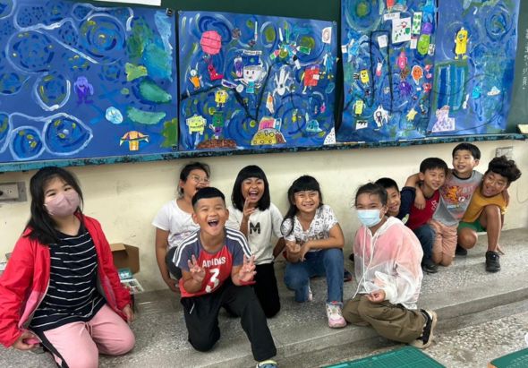 一個教室裡，約十位小學生蹲坐在地板上，開心地擺出各種姿勢和手勢。身後的黑板上貼著多幅藍色主題的兒童繪畫作品，內容包含各式卡通人物和圖案。孩子們穿著校服和便服，臉上帶著笑容，氣氛活潑有趣。