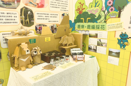 農業、鹿編織花主題展示攤位，黃綠色背景牆上貼滿介紹文字和圖片。展台中央擺放木製動物雕塑、玻璃罐和農產品樣本，展示農業和手工藝相關資訊，呈現教育性的互動展覽。