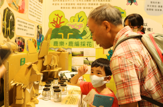 一位長者和小孩在黃色展示攤位前互動。小孩戴著口罩，展示攤位上有鹿茸採花的主題展板、插圖和玻璃罐產品。背景牆面展示相關資訊和教育海報，呈現親子學習的情景。