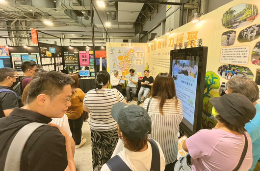 室內展覽會場，眾多參觀者聚集在黃色展板前觀看。展板展示農業相關內容和圖片資訊。左側有書架陳列，天花板可見工業風格的管線和照明設備。參觀者穿著休閒，認真閱讀展示內容。