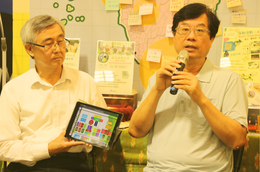 兩位男性在室內進行簡報，右側男性持麥克風講話，左側男性展示平板電腦螢幕。背景牆面貼有黃色便利貼和教育相關海報，桌上放有飲用瓶。場景呈現教育或工作坊的討論氛圍。