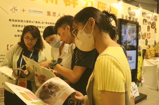 五位年輕參與者在展覽現場聚集，一起查看和討論印刷宣傳冊。背景展示黃色主題的展板和資訊，展覽設有多個展示屏幕，呈現教育推廣或社區活動的互動場景。