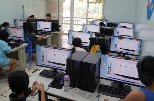 電腦教室內，多名學生坐在各自的工作站前，使用桌上型電腦進行操作。每個工作站配備桌上型主機、顯示器、鍵盤和滑鼠。教室採光良好，窗戶面向戶外綠景。學生專注於螢幕，進行程式設計或資訊相關課程學習。
