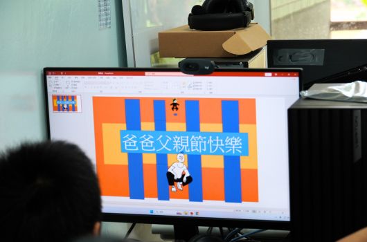 電腦螢幕顯示父親節賀卡設計，背景為橘藍相間的直條紋，中央有卡通人物角色，上方有繁體中文「備備父親節快樂」的祝賀文字，整體色彩鮮艷活潑。