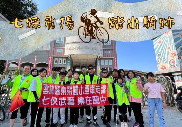 一群穿著螢光黃背心的參與者在「七俠飛揚 踏出騎跡」自行車活動現場合影，手持「豐林尋親族國小單車走讀」和「七俠武尊樂在騎中」的標語，背景為大型戶外展館和自行車雕像。