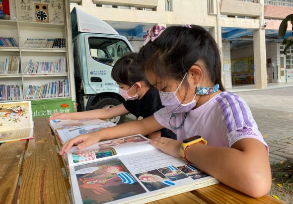 兩名配戴口罩的女孩在戶外閱讀雜誌，坐在木製桌前。背景可見圖書館流動書車、書架和建築物。她們專注地翻閱印有圖片的刊物，享受閱讀時光。