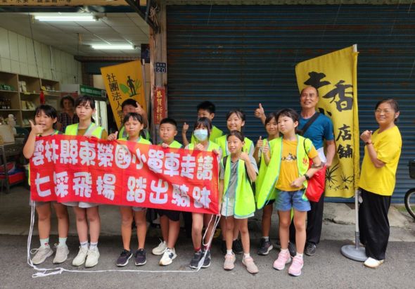 一群穿著黃色和螢光綠背心的學童站在街道上，齊聲舉著紅色布條進行宣傳活動。布條上印有繁體中文標語，後方是店面和黃色招牌，呈現社區參與和公益宣導的場景。
