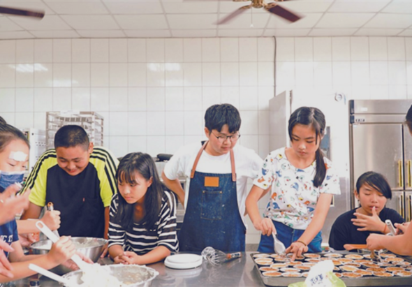 一群小学生在厨房参加烹饪课程，围绕工作台站立，穿着围裙，正在准备食材和烹饪。教室配有不锈钢厨具和天花板风扇，展现了儿童的实践学习活动。