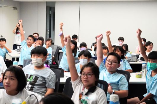 一間教室內，多位穿著淺藍色制服的學生坐在課桌前，舉起雙手做出歡呼慶祝的姿勢。學生們戴著口罩，看起來在參與某項互動式的課堂活動或競賽，氣氛活躍而歡樂。背景可見教室的白牆和門。