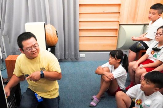 一位穿著黃色T恤的成人音樂教師，坐在椅子上手持吉他，向圍坐在地上的五位兒童進行音樂教學。教室內有灰色窗簾、木製百葉窗和藍色地毯，營造溫暖的學習環境。